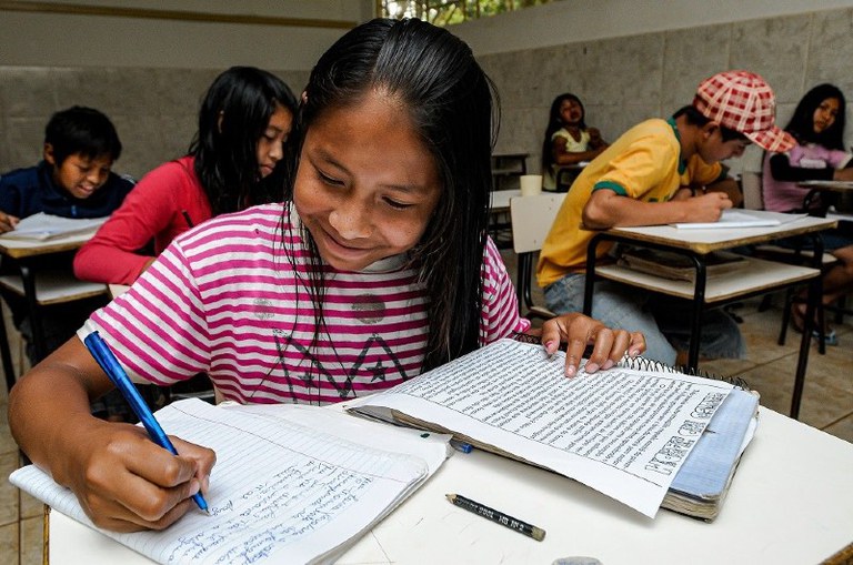 Foto mostra criança indígena sorrindo enquanto escreve em um caderno