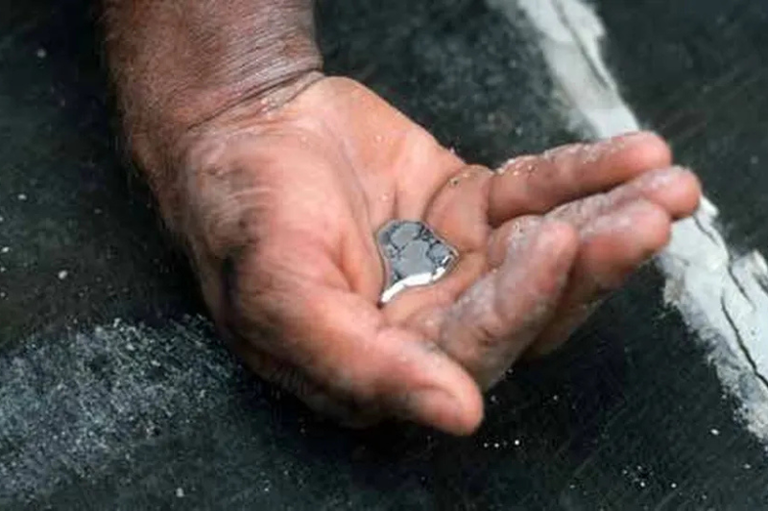 Mão de uma pessoa adulta segurando uma pequena poça prateada de mercúrio líquido, destacando seu brilho e cor. A mão está aberta e sobre uma superfície escura e texturizada.