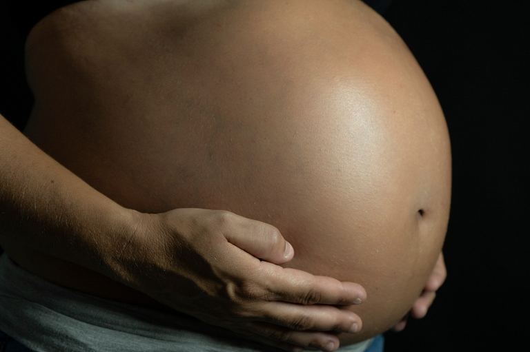 Foto mostra detalhe da barriga de uma mulher negra grávida com as mãos na parte da baixo da barriga; o fundo é escuro