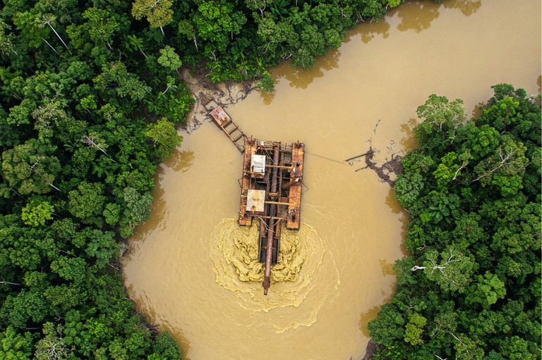 A imagem é uma vista aérea de um rio marrom turvo na Amazônia, cercado por uma densa floresta verde. No centro do rio, uma draga de pequeno porte está em operação, agitando a água e os sedimentos ao seu redor. A perturbação causada pela draga contrasta com a natureza intocada da floresta circundante.