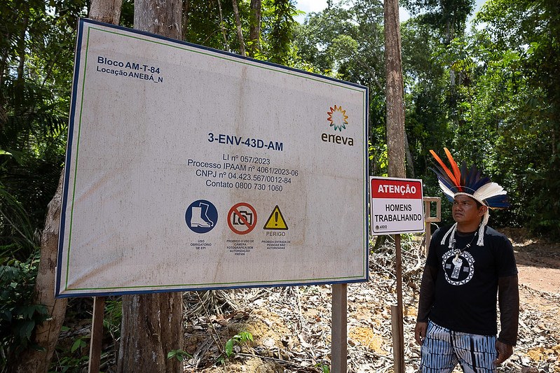 Foto mostra placa grande da empresa Eneva, em área de floresta, com informações sobre licenciamento do empreendimento. Ao lado, uma placa menor diz "Atenção homens trabalhando". Próximo às placas, há um homem moreno com um cocar de penas coloridas