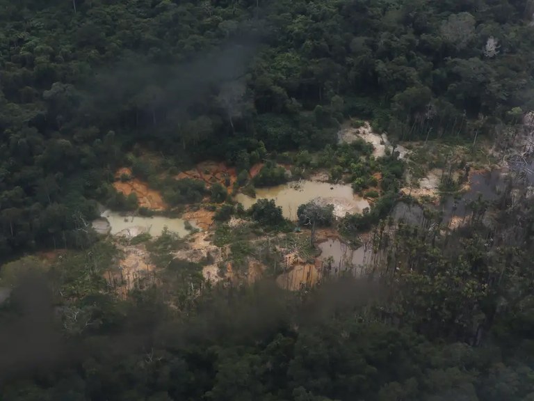 Fotografia aérea mostra abaixo um garimpo ilegal no meio da floresta amazônica. É possível ver o rio, a mata e o estrago causado pelo garimpo, com bancos de terra revolvida nas margens do curso d´água e trechos desmatados.