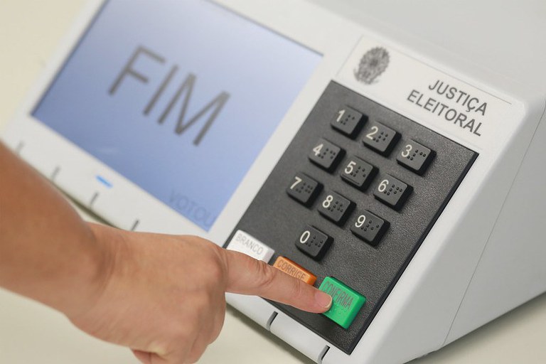 Fotografia em plano aproximado de uma urna eletrônica brasileira. Na tela do aparelho, aparece em destaque a palavra "FIM". No teclado, a mão de uma pessoa pressiona o botão verde "CONFIRMA". Ao lado dele, estão os botões "CORRIGE" (laranja) e "BRANCO" (branco), além do teclado numérico e da inscrição "JUSTIÇA ELEITORAL" no canto superior direito.