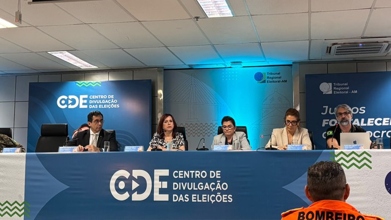 Cinco pessoas sentadas à mesa durante coletiva de imprensa no Centro de Divulgação das Eleições do Tribunal Regional Eleitoral do Amazonas.