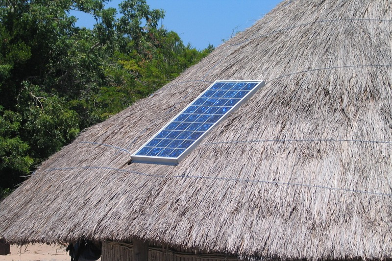 Um painel solar retangular instalado sobre o teto de palha de uma habitação tradicional indígena, cercada por árvores verdes sob um céu azul claro.