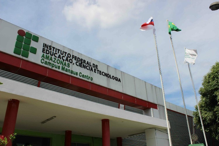 A imagem apresenta a fachada do Instituto Federal de Educação, Ciência e Tecnologia do Amazonas (IFAM), especificamente o Campus Manaus Centro. O prédio possui uma arquitetura moderna com predominância das cores branco, cinza e detalhes em vermelho nas colunas e vigas. No lado esquerdo, destaca-se o logotipo da instituição — uma composição geométrica em tons de verde e um círculo vermelho — acompanhado pelo nome completo do instituto em letras escuras e a identificação do campus em verde. À direita, três mastros exibem as bandeiras do estado do Amazonas, do Brasil e uma bandeira branca institucional, sob um céu claro com poucas nuvens, cercado por copas de árvores que sugerem um ambiente arborizado.