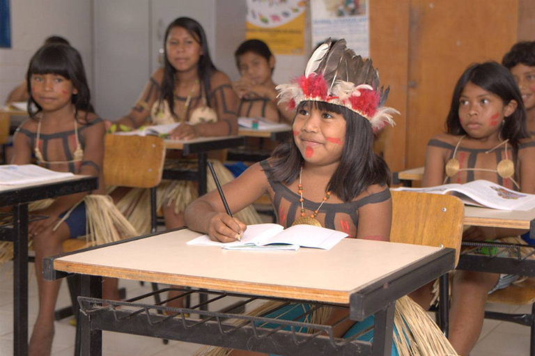 Crianças indígenas sentadas em carteiras escolares em uma sala de aula. A menina em primeiro plano usa um cocar com penas e está escrevendo em um caderno. Todas usam pinturas corporais e vestimentas tradicionais, com saias de palha.
