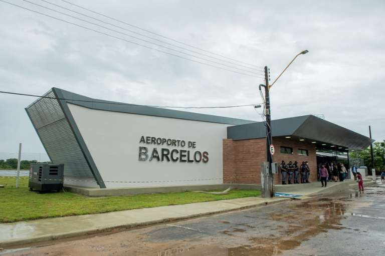Fachada do Aeroporto de Barcelos sob um céu nublado. O terminal é um prédio moderno de cores branca, cinza e marrom, com o nome "Aeroporto de Barcelos" em letras grandes na parede. Na lateral direita, pessoas e policiais circulam pela entrada, e o chão à frente parece molhado pela chuva.