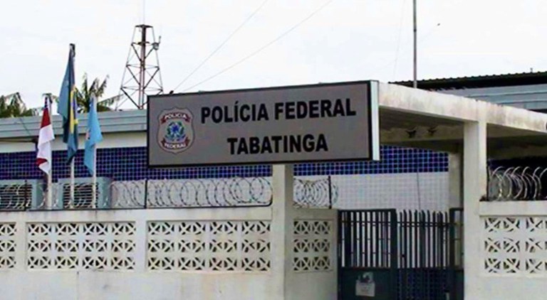 Fachada de um prédio da Polícia Federal em Tabatinga, com uma placa identificando o local, muro com cerca de arame farpado e bandeiras hasteadas na entrada.