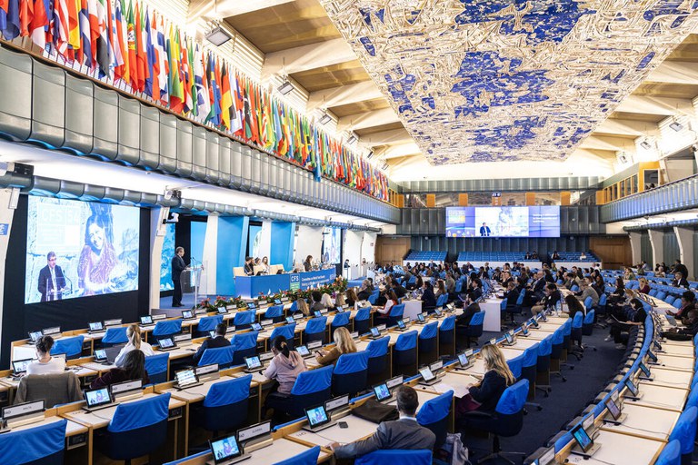 Fotografia num auditório amplo durante a 54ª Sessão do Comité de Segurança Alimentar Mundial da FAO. Dezenas de delegados ocupam bancadas azuis dispostas em arco com ecrãs individuais. Ao fundo, um orador discursa num pódio ao lado da mesa de presidência, sob um ecrã gigante que projeta a sua imagem. O teto ostenta um painel decorativo em tons de azul e branco, e as paredes superiores estão compostas por uma galeria repleta de bandeiras internacionais.