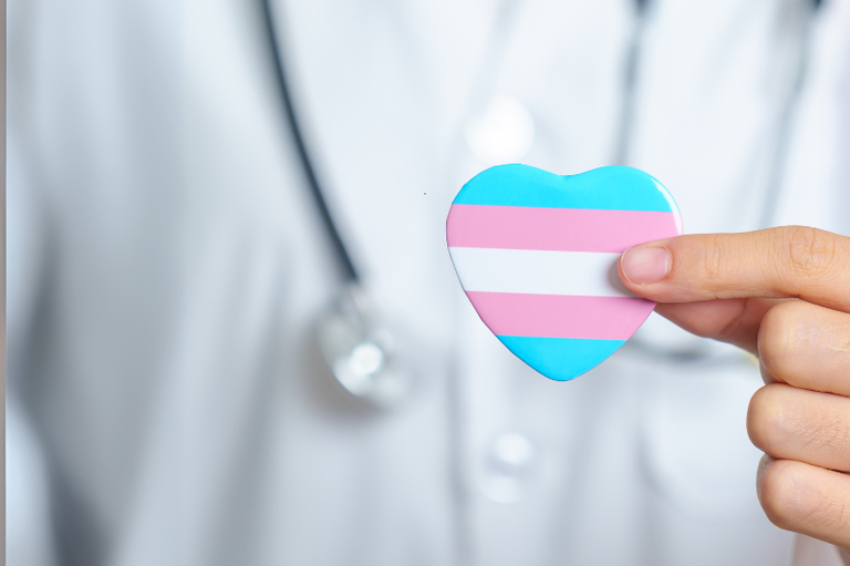 Foto mostra pessoa usando jaleco e com estetoscópio pendurado no pescoço com um pequeno coração nas cores azul, rosa e branco na mão, à frente do corpo, na altura do peito; na foto, não se vê o rosto da pessoa, apenas parte do tronco