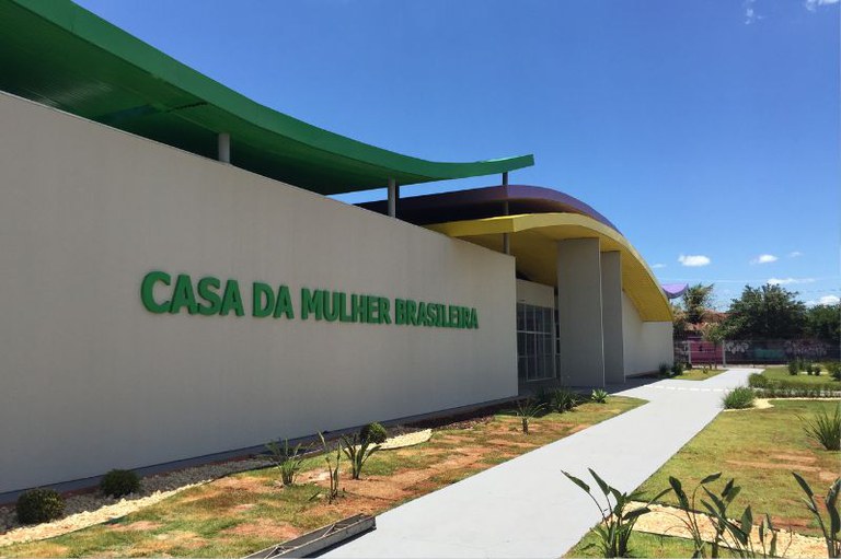 Fotografia em plano médio e perspectiva diagonal da fachada da Casa da Mulher Brasileira em Campo Grande (MS). À esquerda, destaca-se uma grande parede texturizada em tom bege claro com a inscrição em letras verdes e em relevo: "CASA DA MULHER BRASILEIRA". O edifício possui um design moderno, com coberturas curvas sobrepostas nas cores verde, amarela e roxa. À direita, um caminho pavimentado de cor cinza claro corta um jardim gramado com pequenas plantas ornamentais, levando em direção à entrada do prédio, que conta com portas e janelas de vidro sob uma estrutura sustentada por colunas cinzas. Ao fundo, sob um céu azul intenso e poucas nuvens brancas, avistam-se copas de árvores e uma grade delimitando o terreno.