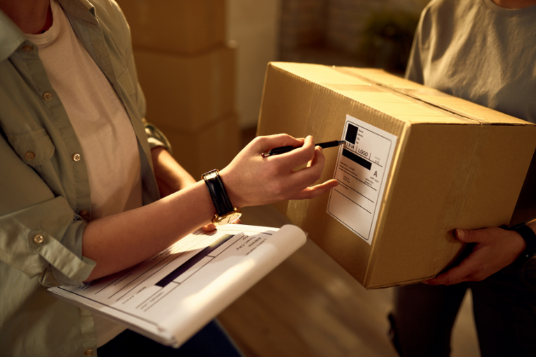 Duas pessoas estão trocando uma caixa de papelão. Uma pessoa, usando um relógio no pulso, aponta para a etiqueta na caixa com uma caneta, enquanto a outra pessoa segura a caixa.