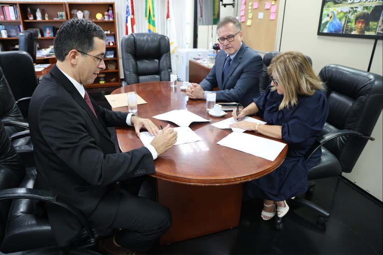 Três pessoas de traje formal estão reunidas em volta de uma mesa oval de madeira em um escritório.