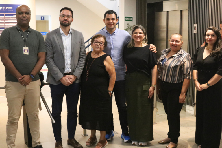 Um grupo de sete pessoas está posando para uma foto em um ambiente interno. Há quatro homens e três mulheres, todos em pé e olhando para a câmera.