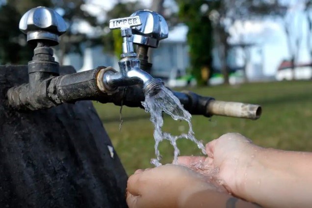 A imagem mostra um par de mãos em concha recebendo água de uma torneira de metal que está conectada a um cano preto. A água jorra com força, salpicando em todas as direções. A torneira parece ser de um local externo, com grama e árvores desfocadas no fundo, sugerindo um ambiente ao ar livre ou rural.