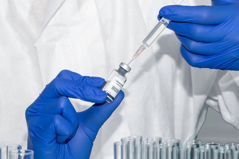 Foto mostra em detalhe um profissional de saúde segurando um frasco de remédio e uma seringa