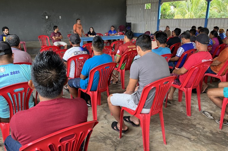 A imagem retrata uma reunião realizada na Comunidade Laranjal, em Nova Olinda do Norte (AM). Um grande grupo de pessoas está sentado em cadeiras de plástico vermelhas. O ambiente é informal, os participantes vestem roupas casuais.