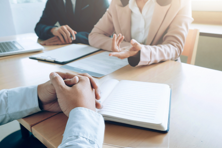 A imagem mostra uma entrevista de emprego ou uma reunião de negócios, com três pessoas sentadas à mesa. As mãos de uma pessoa estão em primeiro plano, sobre um caderno aberto. Ao fundo, uma mulher vestindo um blazer claro gesticula enquanto conversa com outra pessoa, que está parcialmente visível.