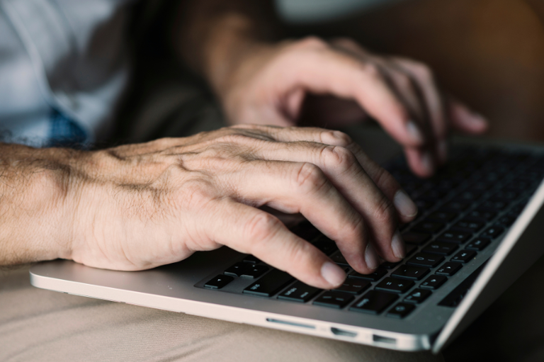 Foto mostra detalhe de homem com as mãos sobre o notebook, apoiado no colo