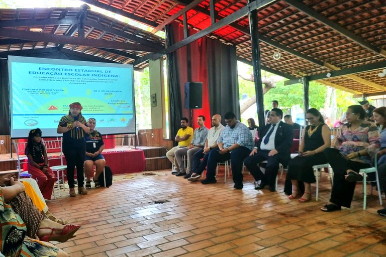 Foto de indígenas e representantes de órgão públicos, entre eles o procurador da República Fernando Soave (de terno), sentados em cadeiras, com três indígenas em frente ao telão de projeção, todos sob uma área coberta com telhado, com uma das indígenas falando ao microfone