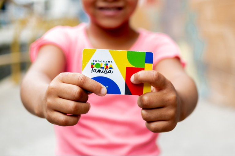 Fotos da mãos de uma criança segurando o cartão colorido do Programa Bolsa Família.