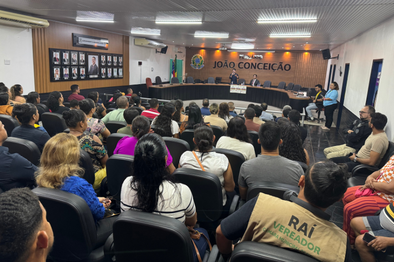 Audiência pública em plenário fechado, com uma grande plateia de pessoas sentadas em cadeiras escuras, vistas por trás. À frente, em uma tribuna elevada, autoridades estão sentados ou em pé, com um painel de madeira ao fundo que exibe "PLENÁRIO JOÃO CONCEIÇÃO".