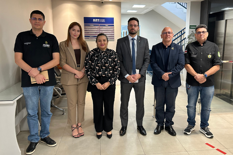 A imagem mostra seis pessoas em pé, lado a lado, no prédio anexo do MPF Amazonas. Há quatro homens e duas mulheres. Os homens vestem roupas casuais de negócios, enquanto as mulheres vestem roupas formais de negócios. Todos estão olhando para a câmera. Ao fundo, há uma parede clara com uma placa azul e branca com letras brancas e uma porta de vidro. O chão é de ladrilhos claros.