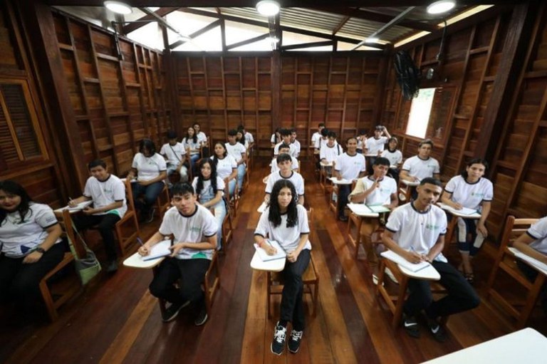 Estudantes de uniforme branco sentados em carteiras individuais dentro de uma sala de aula inteiramente construída em madeira.