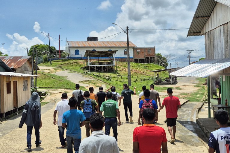 Esta imagem mostra um grupo de aproximadamente 15 jovens e homens caminhando por uma rua de terra e cimento em uma área que parece ser rural ou uma pequena comunidade, possivelmente na Amazônia ou em uma região de clima tropical. Eles estão se afastando da câmera, em direção a um terreno mais alto onde se destacam algumas construções maiores e antigas, incluindo uma com telhado azul e branco e uma estrutura de madeira ao lado, sob um céu parcialmente nublado e ensolarado. A rua é ladeada por casas simples de madeira com telhados de chapa de metal em ambos os lados, e há postes de eletricidade e vegetação exuberante ao redor, sugerindo um ambiente simples e de vida comunitária.