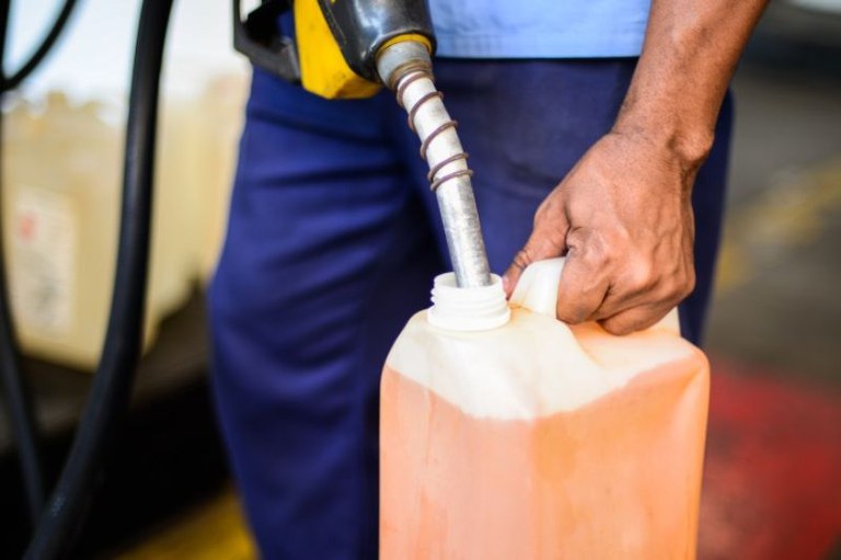 Pessoa vestindo uniforme azul segura um galão plástico enquanto insere o bico de uma bomba de combustível na abertura do recipiente.