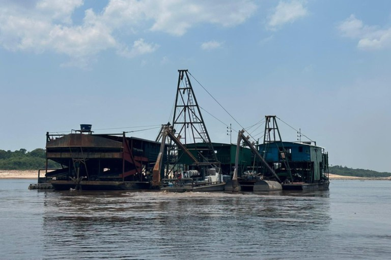 A imagem mostra uma draga grande e industrial, uma espécie de balsa com estruturas metálicas, flutuando em um rio. Ela é usada para mineração e possui guindastes e tubulações visíveis. A draga está no centro do rio, com a mata nativa e a margem visíveis ao fundo, sob um céu parcialmente nublado.