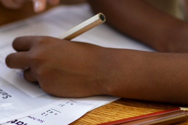 Imagem mostra em detalhe a mão de uma criança escrevendo com um lápis branco em um caderno