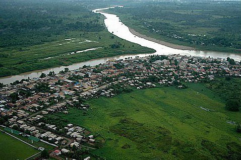 A imagem mostra uma vista aérea de uma cidade às margens de um rio sinuoso, cercada por áreas verdes e vegetação densa. As casas estão concentradas próximas ao rio, enquanto o restante da paisagem é majoritariamente rural e natural.