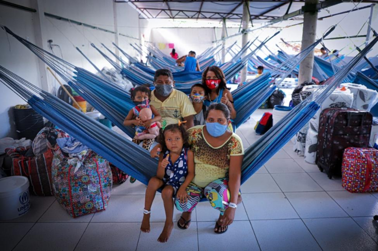 Família usando máscaras de proteção posa em um abrigo repleto de redes azuis armadas e pertences pessoais.
