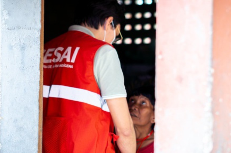 Foto mostra um profissional de saúde da Sesai com um colete vermelho. Há uma indígena sentada.