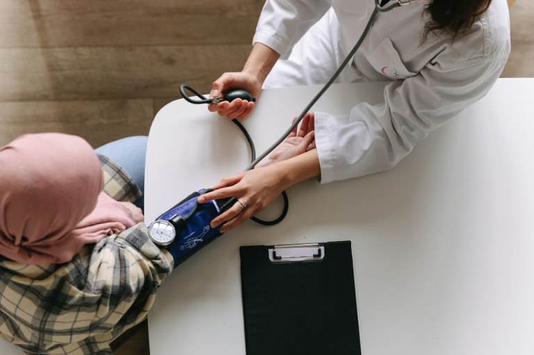 Foto mostra profissional da saúde medindo a pressão de uma paciente com câncer. Ela usa um lenço rosa na cabeça.