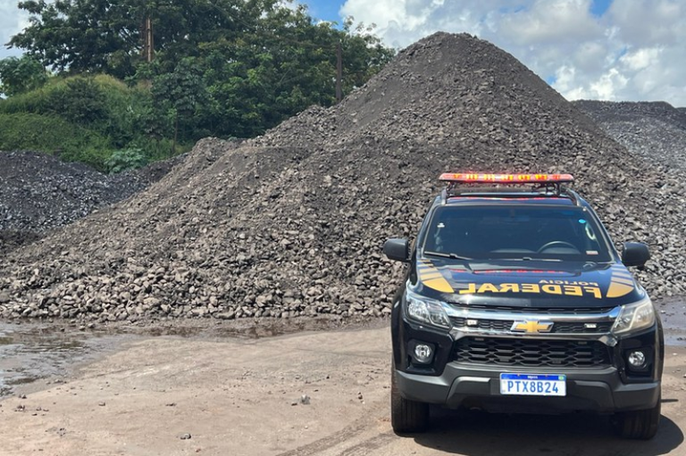 Foto mostra um monte de minério apreendido em operação policial, acumulado em ambiente aberto, tendo, à frente, uma viatura da Polícia Federal