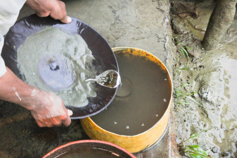 Fotografia dos braços de um homem segurando uma bandeja contendo lama e mercúrio, utilizado para garimpo ilegal de ouro, que estão sendo derramados em um balde contendo água com lama e um recipiente de vidro