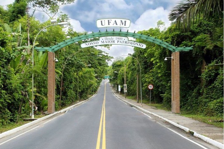 Pórtico de entrada com a sigla "UFAM" e a frase "Nosso Maior Patrimônio" sobre uma estrada asfaltada cercada por vegetação densa.