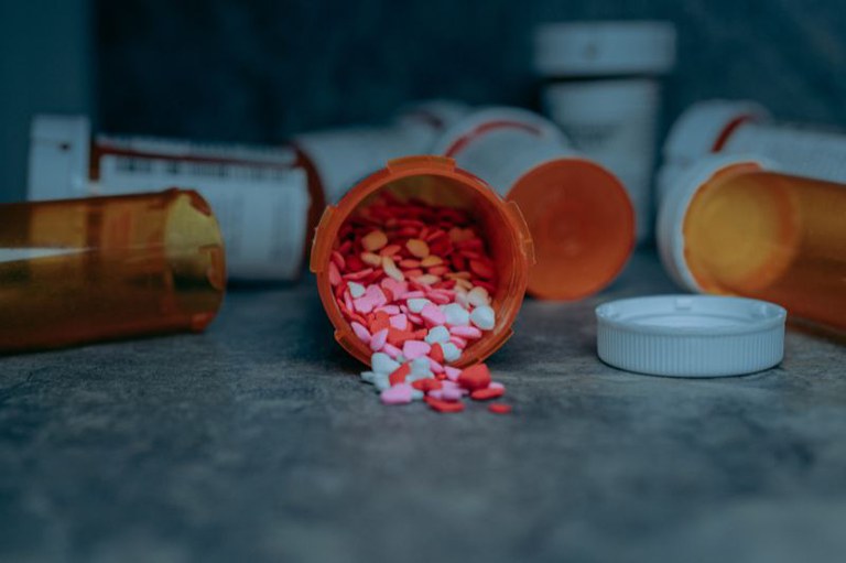 Foto mostra diversos frascos de medicamentos deitados em uma superfície cinza, sendo que um deles está aberto e os comprimidos coloridos estão esparramados