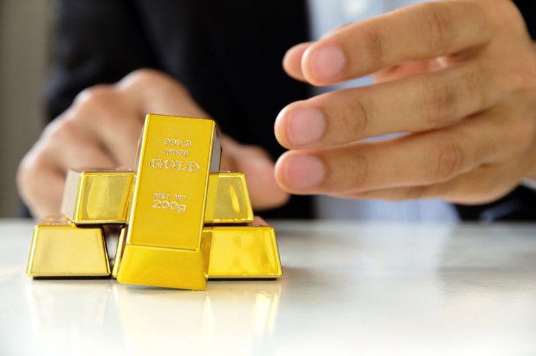 Pequena pilha de barras de ouro brilhantes sobre uma superfície clara e reflexiva, com uma barra em destaque na vertical mostrando a gravação "999.9 FINE GOLD NET WT 200g". Ao fundo, as mãos desfocadas de uma pessoa de terno se aproximam do ouro.
