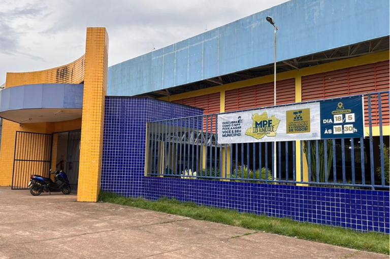A imagem mostra a fachada de uma escola, apresentando paredes em tons de azul e colunas revestidas por pastilhas amarelas. Fixada à grade metálica azul, destaca-se uma faixa informativa do programa "MPF na Comunidade", indicando um evento no estado do Amazonas agendado para o dia 18 de maio, às 18h30.