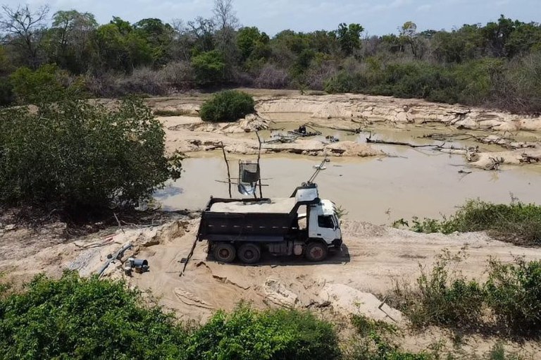 Foto mostra um caminhão, com uma estrutura que deposita areia em sua caçamba. Ele está sobre uma área com muita areia, e também alguma vegetação ao redor.