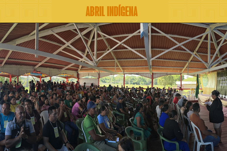 Fotografia mostra um enorme galpão, lotado com mais de 150 pessoas que aguardam o início de um evento. Ao fundo, é possível ver um rio e a floresta na linha do horizonte.