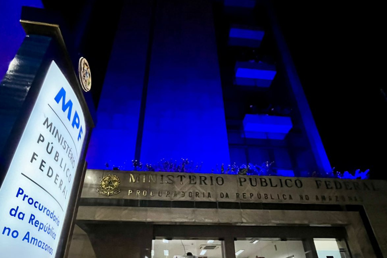 A imagem mostra a entrada principal do prédio do Ministério Público Federal no Amazonas. O prédio está iluminado com luzes azuis, em alusão ao  Dia Mundial Contra o Tráfico de Pessoas, celebrado no dia 30 deste mês. Na parte superior da imagem, vê-se a inscrição "MINISTÉRIO PÚBLICO FEDERAL" e logo abaixo, "PROCURADORIA DA REPÚBLICA NO AMAZONAS" gravadas em uma faixa metálica acima das portas de entrada. À esquerda, há um totem iluminado com o logotipo do MPF (Ministério Público Federal) e a identificação do local: "Procuradoria da República no Amazonas". A entrada do edifício é acessada por uma escada com corrimão de aço inox e portas de vidro automáticas, através das quais é possível ver o interior iluminado, com recepção e áreas de atendimento.
