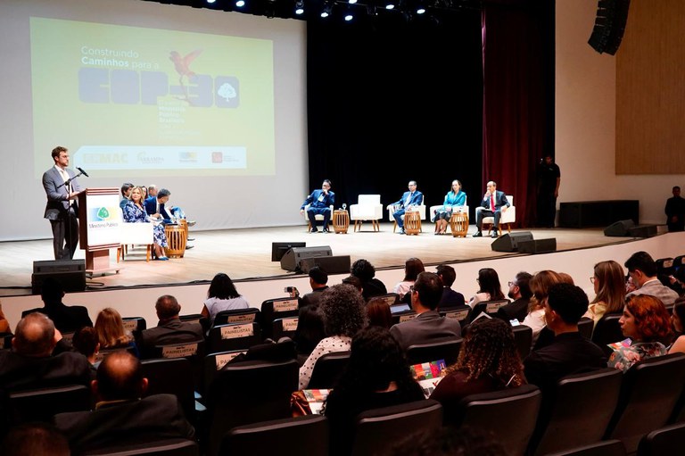 Foto mostra homem de terno cinza claro em frente a púlpito com microfone; ao fundo, há uma projeção com a expressão Construindo caminhos COP30