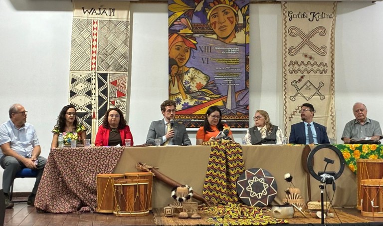 foto mostra mesa de abertura do Congresso Socioambiental, procurador da República João Pedro está ao centro, de terno cinza e microfone em mãos. Há outras sete pessoas sentadas compondo a mesa.