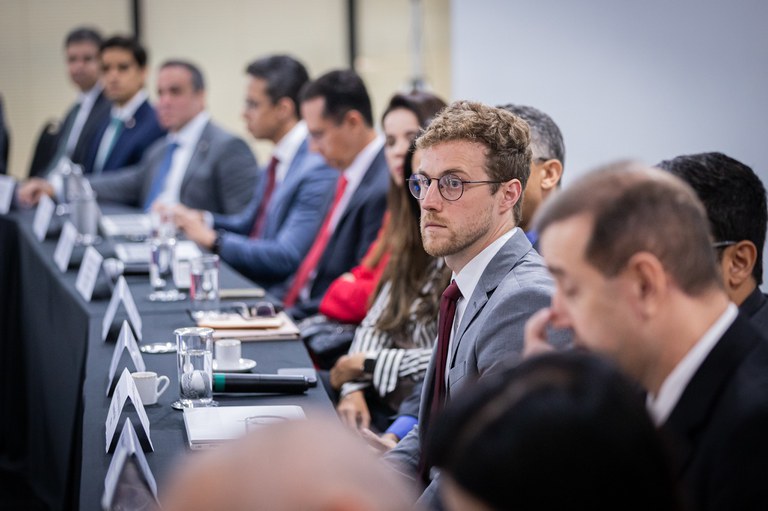 Homem branco, de olhos claros, usando terno, em primeiro plano. Ele compçõe, ao lado de outros homens vestidos formalmente, uma mesa redonda em sala de reuniões