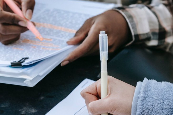 A imagem mostra mãos de duas pessoas em uma atividade de estudo. Uma delas utiliza um marca-texto cor-de-rosa para destacar trechos em uma folha com anotações, e a outra segura uma caneta. Ambas estão com papéis e documentos à frente.